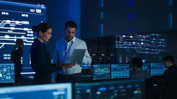 A man and a woman looking at a document in a room full of computer screens.