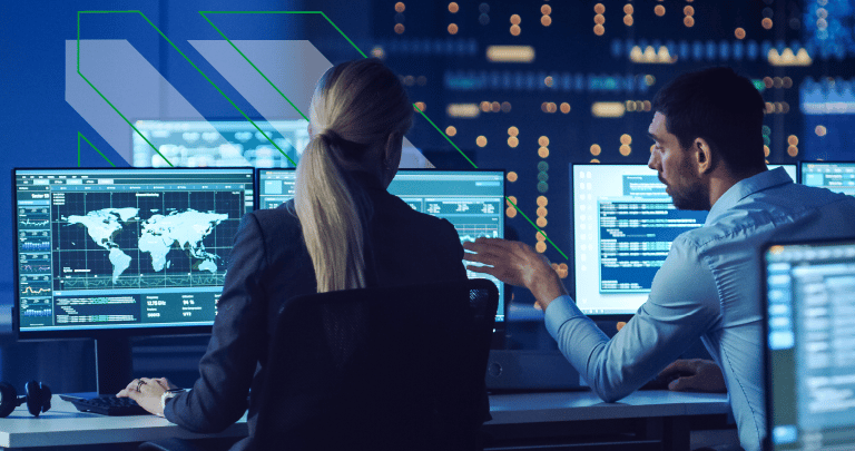 Two cyber security professionals sat at their desk in a network control room, discussing work and monitoring computers.