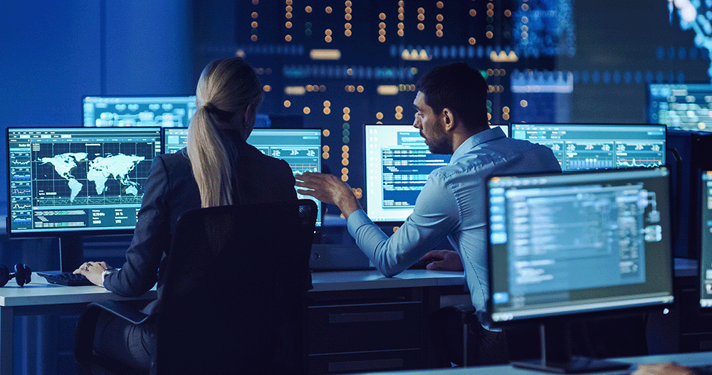 Two IT professionals discussing work at their desks, whilst monitoring computer screens.