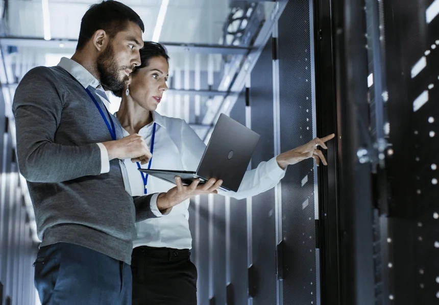 Male IT technician holding laptop next to female IT technician who is pointing at server in data center.