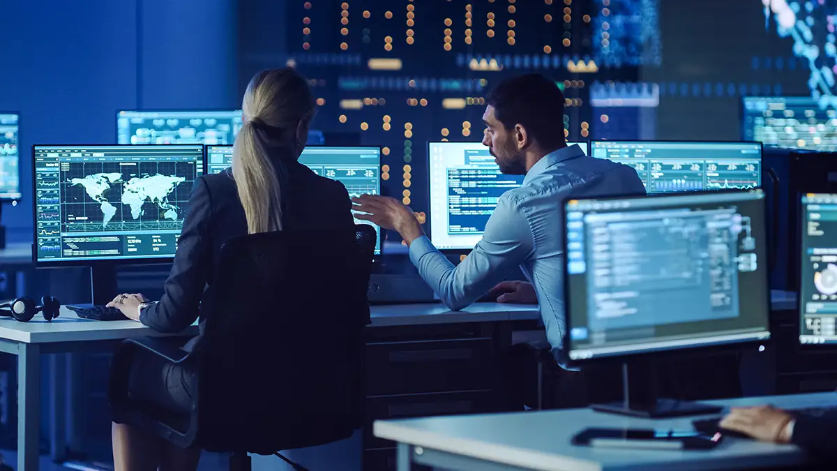 Two cyber security professionals discussing security and compliance posture at their desks, whilst monitoring computers.