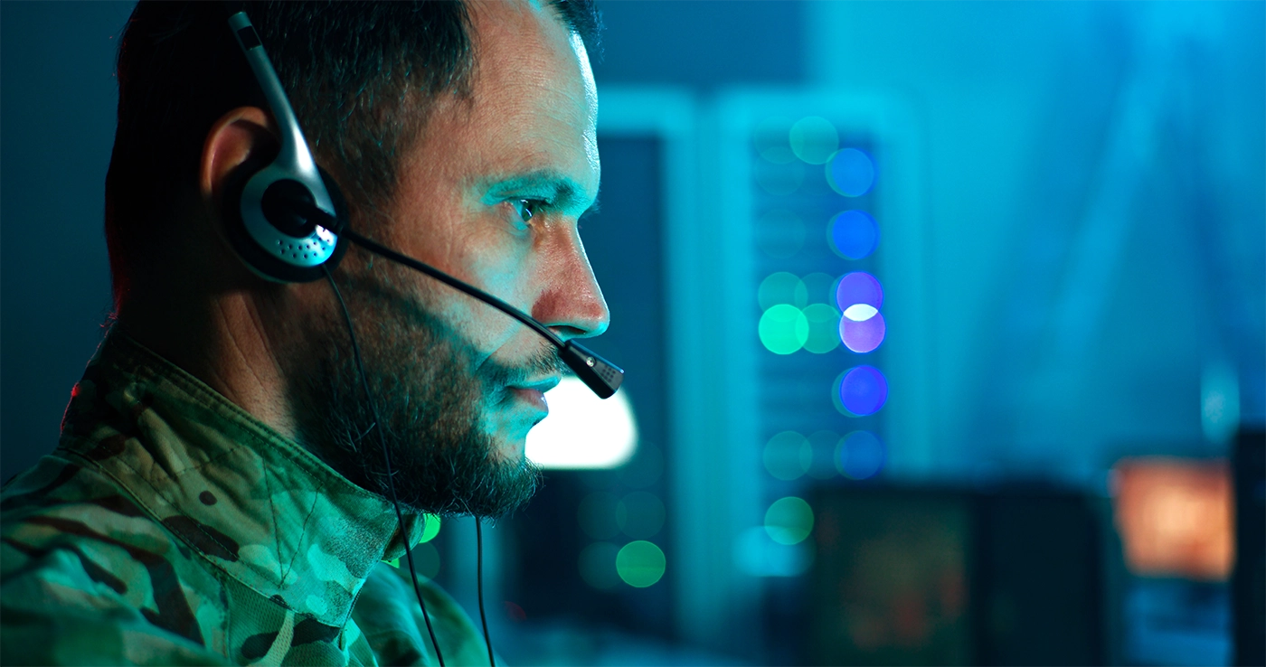 Military cyber security professional speaking on headset and using computer while working in network control center.