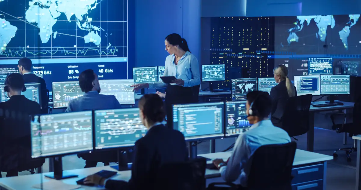 Senior Cyber Security Professional holding Tablet and discussing work with Engineer in telecommunications control monitoring room, surrounded by team members.